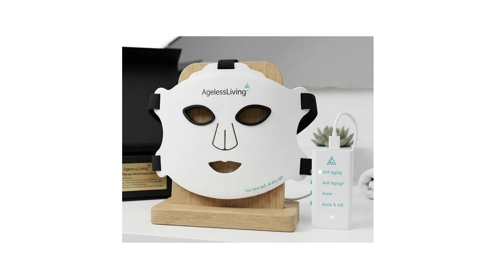 The Ageless Living Red Light and Acne Therapy Mask on its wooden stand beside its controller