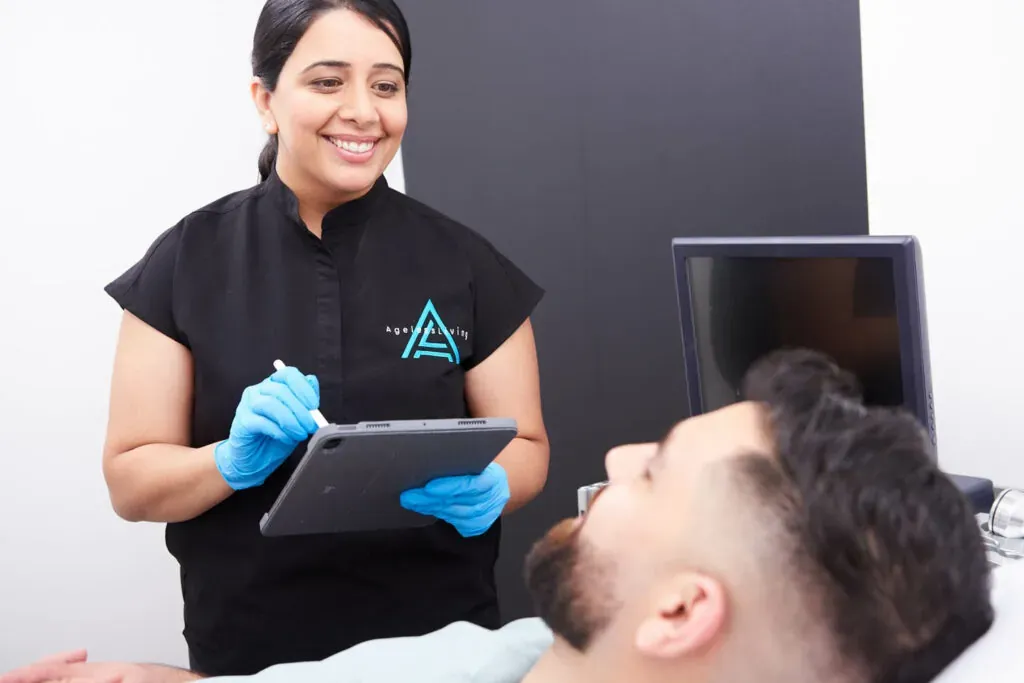 An Ageless Living medical aesthetician in gloves reviewing a treatment plan on a tablet beside a patient on the treatment bed