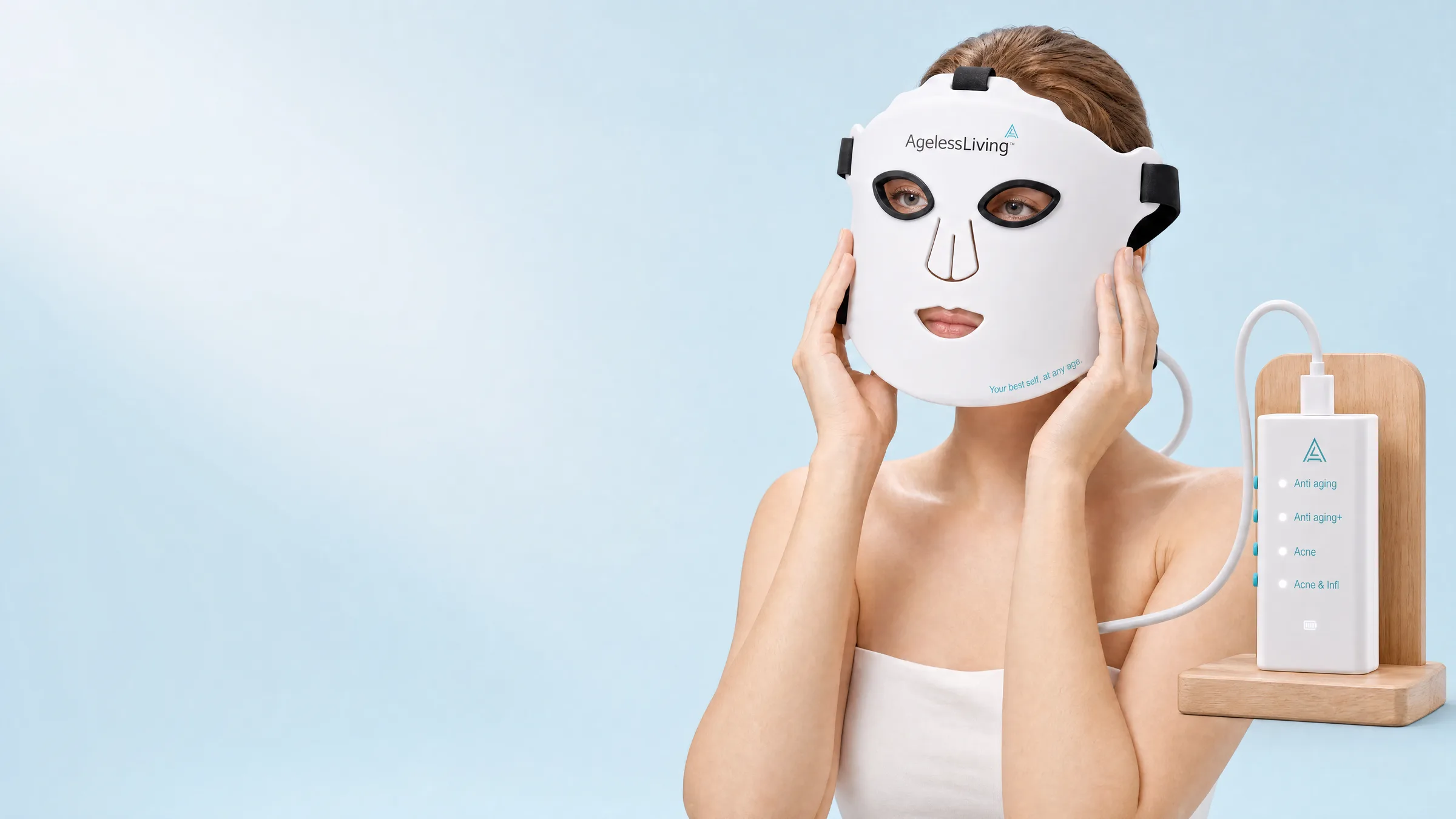 A woman holding an Ageless Living LED face mask to her face beside its controller