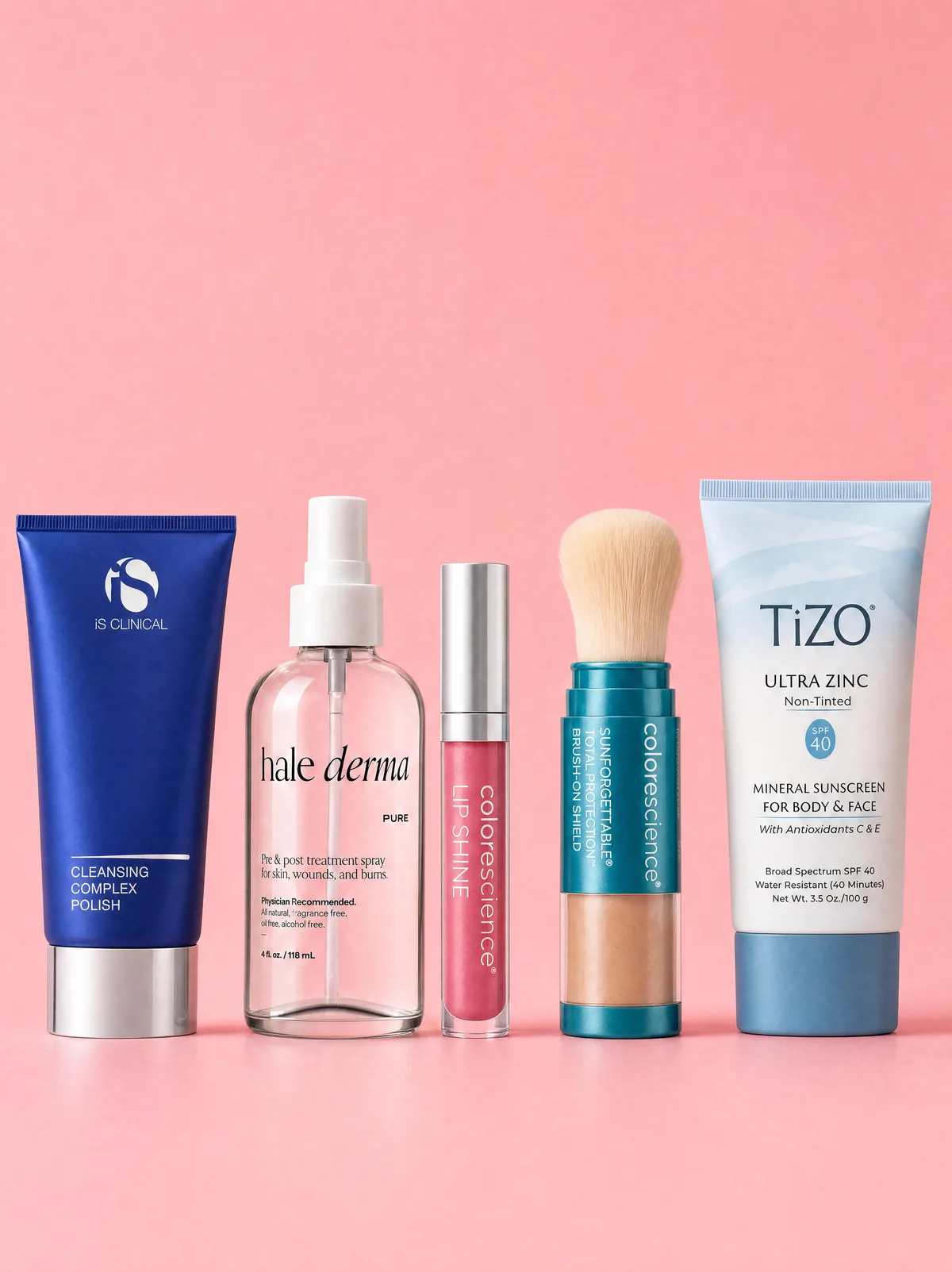 Five clinic skincare products in a row on pink: a cleansing polish, a treatment spray, a lip shine, a brush-on mineral shield and a zinc sunscreen