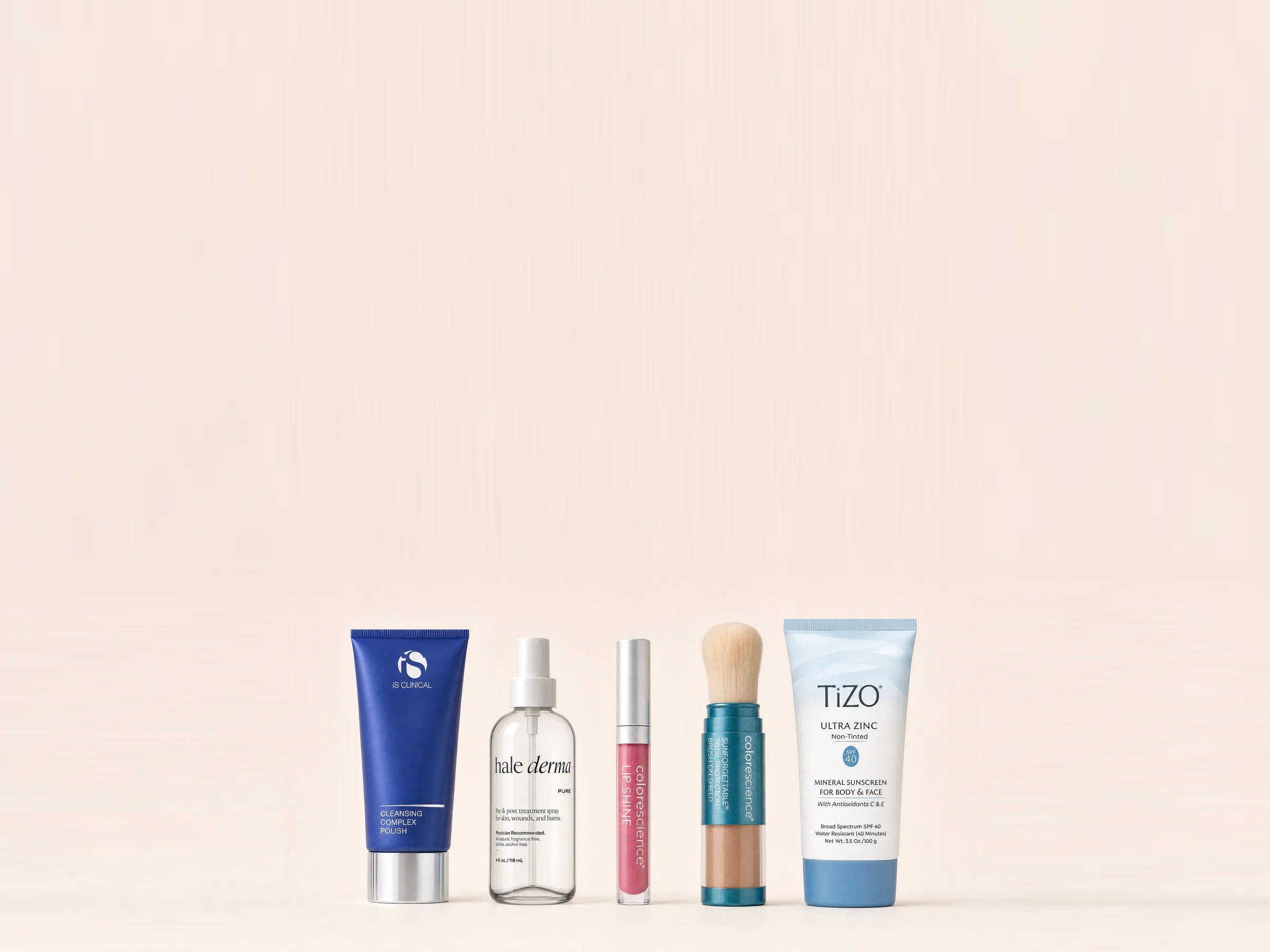 Five clinic skincare products in a row on cream: a cleansing polish, a treatment spray, a lip shine, a brush-on mineral shield and a zinc sunscreen