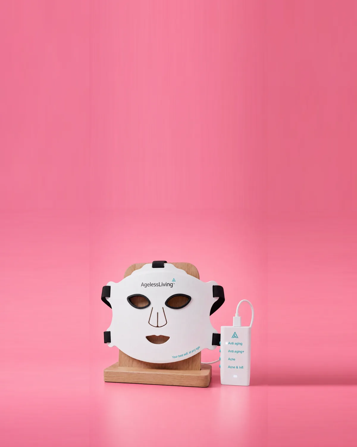 An Ageless Living LED mask on a wooden stand beside its controller, on pink
